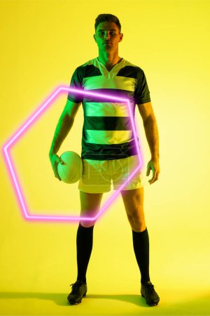 Photo for Serious male caucasian rugby player with ball by illuminated triangle standing on yellow background. Copy space, composite, sport, shape, competition, confident, playing, match and abstract concept. - Royalty Free Image