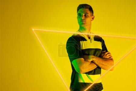 Photo for Male caucasian rugby player with arms crossed standing by illuminated triangle on yellow background. Copy space, composite, sport, shape, competition, playing, match and abstract concept. - Royalty Free Image