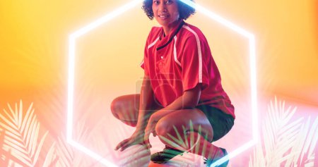 Photo for Biracial smiling female rugby player crouching with ball over illuminated hexagon and plants. Copy space, composite, sport, competition, shape, nature, playing, match and abstract concept. - Royalty Free Image