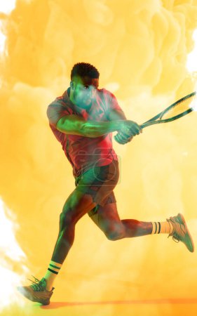 Photo for African american male tennis player hitting with racket against smoky yellow background. Copy space, composite, sport, competition, playing, match and abstract concept. - Royalty Free Image