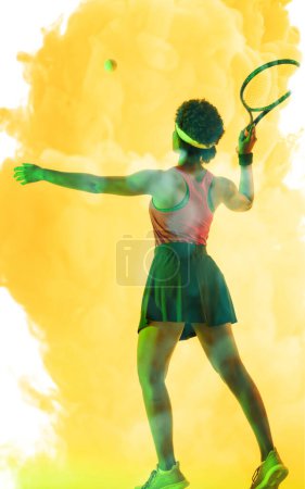 Photo for Rear view of african american female player hitting ball with tennis racket on smoky background. Copy space, yellow, composite, sport, competition, playing, match and abstract concept. - Royalty Free Image