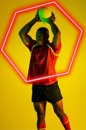Photo for African american male rugby player throwing ball by illuminated hexagon over yellow background. Copy space, composite, sport, competition, shape, playing, match and abstract concept. - Royalty Free Image