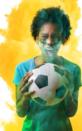 Photo for African america female fan with brazilian flag's face paint and ball screaming on smoky background. Composite, sport, competition, soccer, support, cheering, mouth open, excited, match, illuminated. - Royalty Free Image
