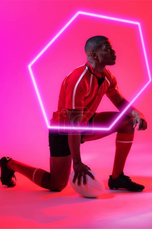 Téléchargez les photos : Joueur afro-américain avec ballon de rugby agenouillé par hexagone illuminé sur fond rose. Espace de copie, composite, sport, compétition, forme, jeu, match et concept abstrait. - en image libre de droit
