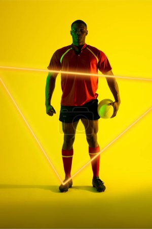 Téléchargez les photos : Joueur afro-américain confiant avec balle de rugby par triangle éclairé sur fond jaune. Espace de copie, composite, sport, compétition, forme, jeu, match et concept abstrait. - en image libre de droit