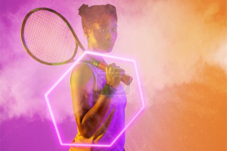 Téléchargez les photos : Une joueuse de tennis afro-américaine confiante tenant une raquette au milieu de la fumée par un hexagone lumineux. Espace de copie, composite, sport, forme, compétition, jeu, match et concept abstrait. - en image libre de droit