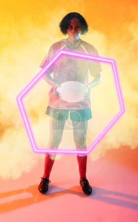 Téléchargez les photos : Joueuse de rugby biraciale confiante avec balle debout au milieu de la fumée sur hexagone éclairé. Copier espace, sport, compétition, forme, fumée, jeu, match et concept abstrait. - en image libre de droit