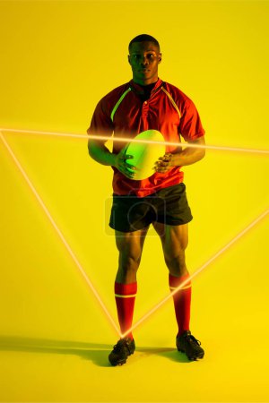 Foto de Confiado jugador de rugby afroamericano masculino con pelota por triángulo iluminado sobre fondo amarillo. Copiar espacio, compuesto, deporte, competencia, forma, juego, partido y concepto abstracto. - Imagen libre de derechos