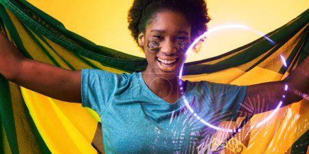 Photo for Cheerful african american female fan with brazilian flag and face paint by circle and plants. Composite, sport, competition, soccer, shape, support, nature, match, illuminated and patriotism. - Royalty Free Image