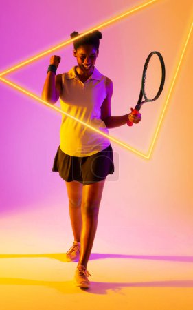Téléchargez les photos : Heureuse joueuse afro-américaine de tennis tenant la raquette et secouant le poing par un triangle lumineux. Espace de copie, composite, forme, sport de célébration, compétition, vainqueur, match et concept abstrait. - en image libre de droit