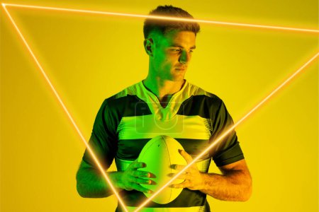 Photo for Caucasian male rugby player with ball looking away by illuminated triangle over yellow background. Copy space, composite, sport, shape, competition, playing, match and abstract concept. - Royalty Free Image
