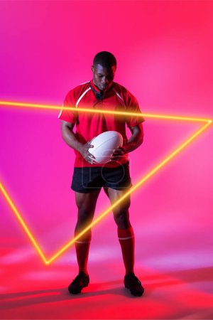 Photo for Confident african american male rugby player with ball by illuminated triangle on pink background. Copy space, composite, sport, competition, shape, playing, match and abstract concept. - Royalty Free Image