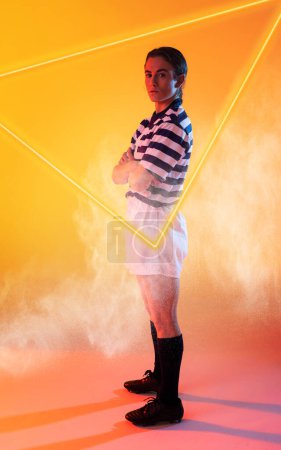 Foto de Confiada jugadora de rugby caucásica con los brazos cruzados de pie por triángulo iluminado. Copiar espacio, compuesto, deporte, competición, forma, humo, partido y concepto abstracto. - Imagen libre de derechos