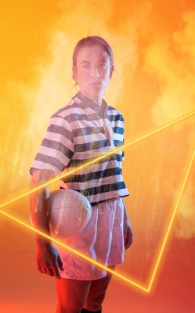 Téléchargez les photos : Joueuse caucasienne confiante tenant le ballon et se tenant près d'un triangle illuminé au milieu de la fumée. Espace de copie, composite, sport, compétition, forme, fumée, jeu, match et rugby concept. - en image libre de droit