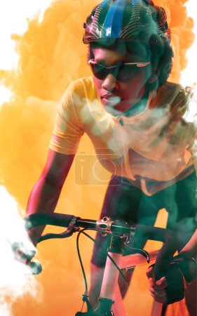 Téléchargez les photos : Confident african american female cyclist wearing glasses and helmet riding bike on smoky background. Copy space, yellow, composite, sport, cycling, racing, competition and abstract concept. - en image libre de droit