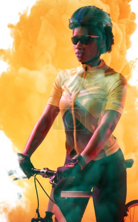 Téléchargez les photos : Confident african american cyclist wearing helmet and glasses standing with bike on smoky background. Copy space, composite, yellow, sport, cycling, racing, competition and abstract concept. - en image libre de droit