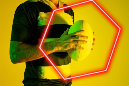 Téléchargez les photos : Illuminated hexagon over midsection of caucasian male player holding rugby ball on yellow background. Copy space, composite, sport, competition, shape, playing, match, hand and abstract concept. - en image libre de droit