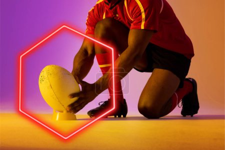 Téléchargez les photos : Illuminated hexagon over low section of african american male rugby player placing ball on stand. Copy space, composite, sport, competition, shape, playing, hand, red, match and abstract concept. - en image libre de droit