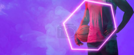 Téléchargez les photos : Midsection of biracial basketball player with ball standing by hexagon over smoky background. Blue, composite, copy space, hand, sport, competition, illustration, illuminated, shape and abstract. - en image libre de droit