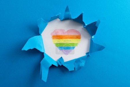 Téléchargez les photos : Close up of torn paper and rainbow coloured heart with copy space on blue background. Pride month, equality, lgbt and human rights concept. - en image libre de droit