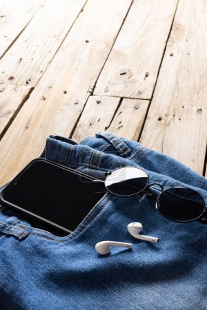 Photo for Vertical image of blue jeans with smartphone, earphones and glasses lying on wooden surface. Clothes, fashion, design, fabrics, materials and shopping concept. - Royalty Free Image