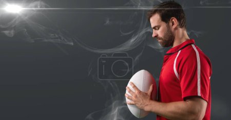 Foto de Jugador de rugby masculino caucásico sosteniendo la pelota contra el efecto de humo y la mancha de luz sobre fondo gris. torneo deportivo y concepto de competición - Imagen libre de derechos