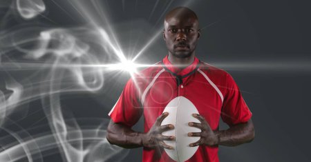 Photo for African american male rugby player holding ball against smoke and light spot on grey background. sports tournament and competition concept - Royalty Free Image
