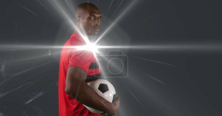 Téléchargez les photos : African american male soccer player holding football against smoke and light spot on grey background. sports tournament and competition concept - en image libre de droit