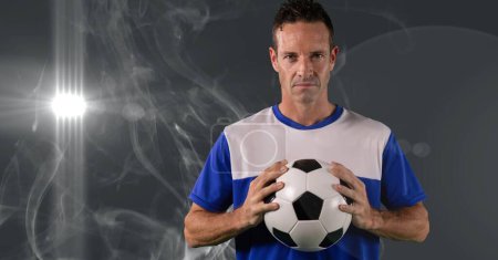 Photo for Caucasian male soccer player holding football against smoke effect and light spot on grey background. sports tournament and competition concept - Royalty Free Image