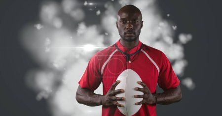 Téléchargez les photos : Portrait of african american male rugby player holding ball against light spot on grey background. sports tournament and competition concept - en image libre de droit