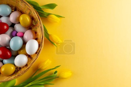 Photo for Image of multi coloured easter eggs in basket with yellow tulips and copy space on yellow background. Easter, religion, tradition and celebration concept. - Royalty Free Image