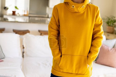 Téléchargez les photos : Milieu de la femme biraciale portant un sweat-shirt jaune avec espace de copie dans le salon. Mode, design, couleur et concept de vêtements. - en image libre de droit