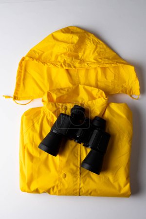 Téléchargez les photos : Gros plan de manteau imperméable jaune et jumelles sur fond blanc avec espace de copie. Mois national de camping, équipement et concept de célébration. - en image libre de droit