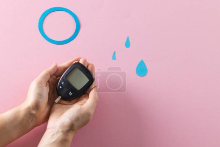 Photo for Hands of caucasian woman holding glucometer over blue drops and ring on pink background, copy space. Blood sugar, diabetes and health awareness. - Royalty Free Image