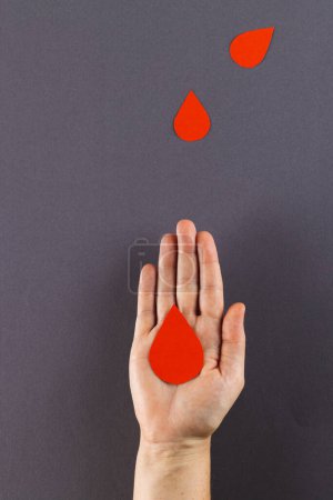 Téléchargez les photos : Main de femme caucasienne tenant une goutte de sang, avec des gouttes de sang sur fond gris, espace de copie. Don de sang, médicaments et soins de santé. - en image libre de droit