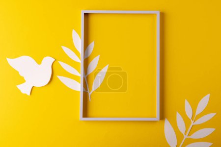 Photo for High angle view of white frame, white dove and leaves with copy space on yellow background. Peace and anti war movement concept. - Royalty Free Image