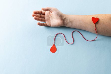 Foto de Brazo de mujer caucásica con corazón y cuerda roja con gota de sangre, sobre fondo azul. Donación de sangre, medicamentos y asistencia sanitaria. - Imagen libre de derechos
