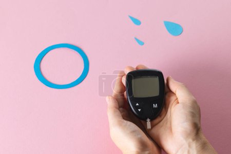 Photo for Hands of caucasian woman holding glucometer over blue drops and ring on pink background. Blood sugar, diabetes and health awareness. - Royalty Free Image