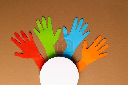 Photo for Paper cut out of multi coloured hands and white circle with copy space on brown background. Humanitarian aid, people, help and human concept. - Royalty Free Image