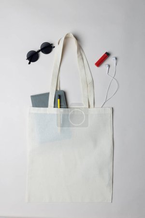 Photo for High angle view of white canvas bag with notebook, sunglasses, earphones and lighter, copy space. Bags and fashion concept. - Royalty Free Image
