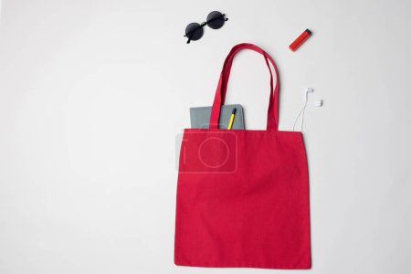 Foto de Vista de ángulo alto de la bolsa de lona roja con portátil, gafas de sol, auriculares y encendedor, espacio para copiar. Bolsas y concepto de moda. - Imagen libre de derechos