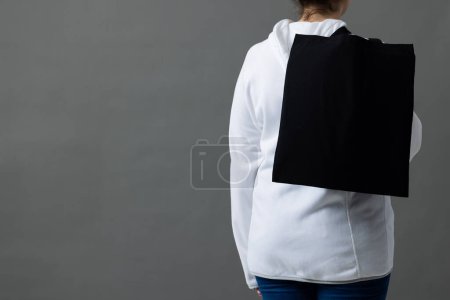 Foto de Mujer caucásica sosteniendo sobre el hombro bolsa de lona negra con espacio de copia sobre fondo gris. Bolsas y concepto de moda. - Imagen libre de derechos