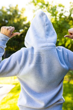 Téléchargez les photos : Vue arrière de la femme caucasienne pointant du doigt et portant un sweat à capuche gris sur le parc vert par une journée ensoleillée. Vêtements, matériel et modèle. - en image libre de droit