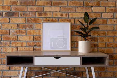 Photo for Wood empty frame with copy space and plant in pot on desk against brick wall. Mock up frame template, interior design and decoration. - Royalty Free Image