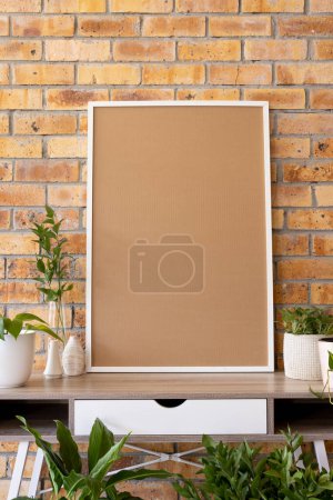 Photo for Vertical of empty white frame with copy space and plants in pots on desk against brick wall. Mock up frame template, interior design and decoration. - Royalty Free Image