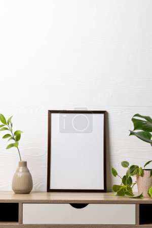 Photo for Vertical of black empty frame with copy space and plants in pots on table against white wall. Mock up frame template, interior design and decoration. - Royalty Free Image
