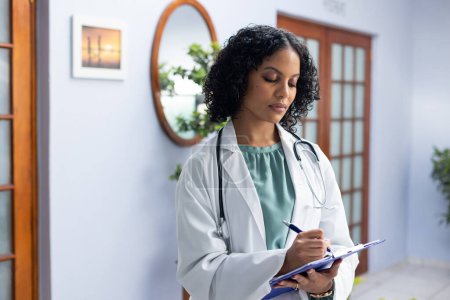 Photo for Biracial female doctor wearing stethoscope holding clipboard and taking notes at doctor's office. Hospital, medicine and healthcare. - Royalty Free Image