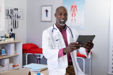Téléchargez les photos : Portrait d'un heureux médecin afro-américain portant un stéthoscope à l'aide d'une tablette au cabinet du médecin. Hôpital, médecine, santé et communication. - en image libre de droit
