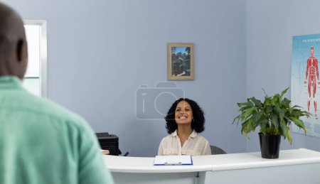 Photo for Biracial medical receptionist sitting at reception desk and smiling at african american patient. Hospital, medicine and healthcare. - Royalty Free Image