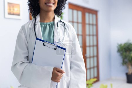 Photo for Biracial female doctor wearing stethoscope holding clipboard and smiling at doctor's office. Hospital, medicine and healthcare. - Royalty Free Image
